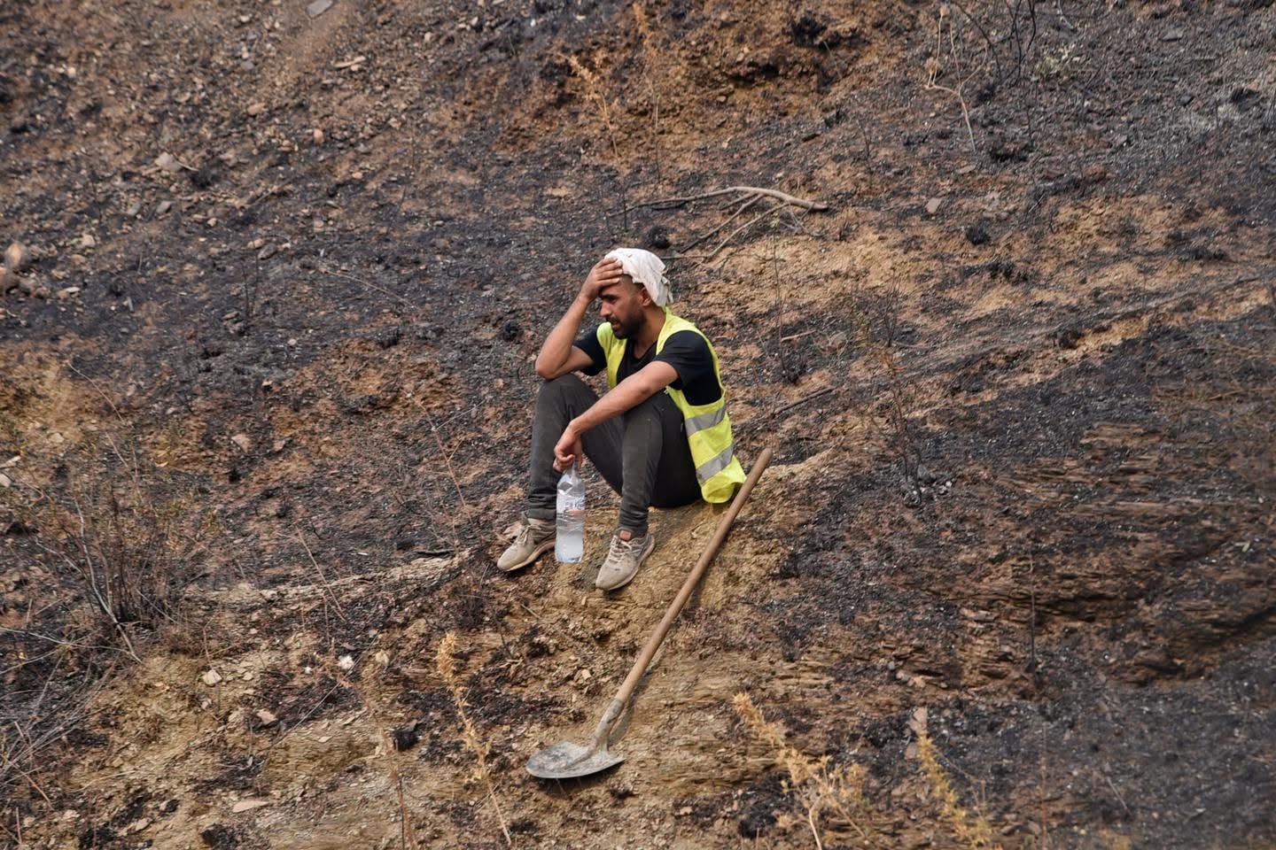 Un homme assis sur une colline brûlée, près d’une pelle et d’une bouteille d’eau.